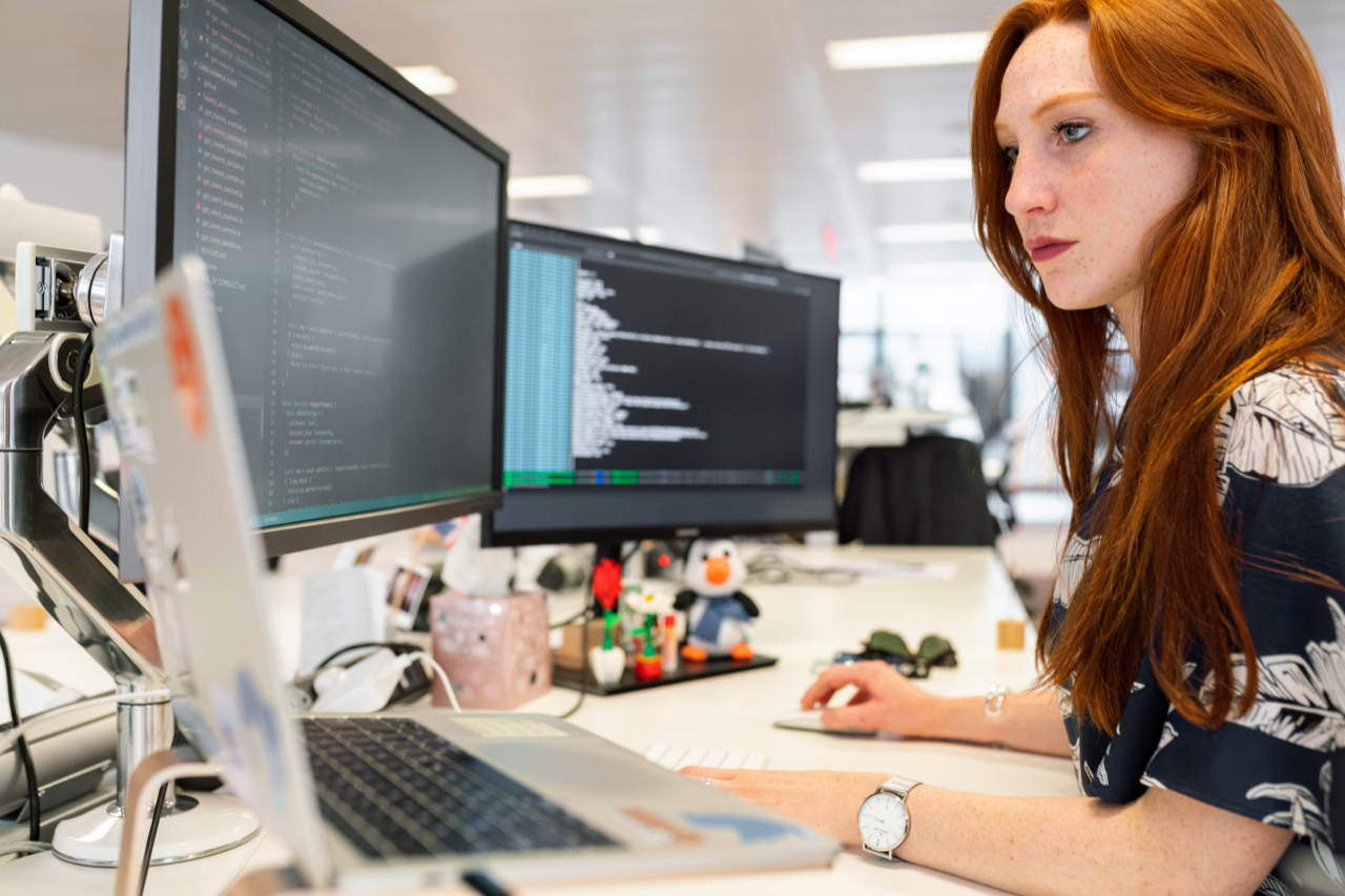 Red-headed women working at a desk with a laptop and two monitors in front of her